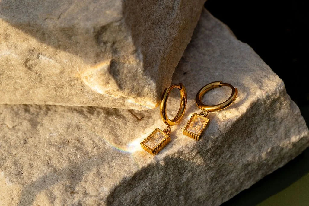 A pair of gold hoop earrings with rectangular drop pendants featuring a textured gold border and a clear cubic zirconia center, resting on a textured beige rock under warm sunlight. Features the Rectangle Glister Huggies 