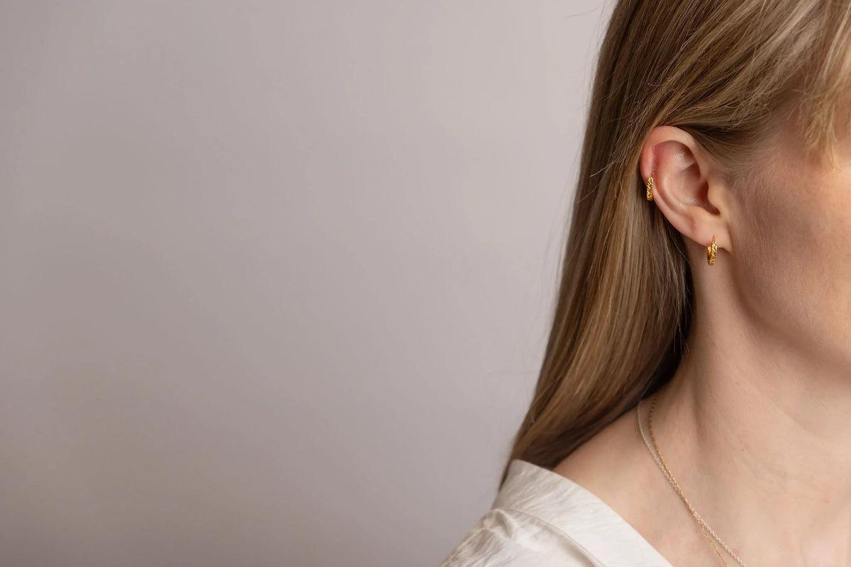 A close-up side profile of a woman wearing small gold stack hoop earrings and a delicate gold necklace, set against a plain light grey background. One earring is the helix and one is in the lobe. 