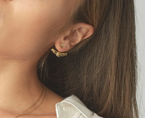 A close-up of a model's ear wearing a gold geometric fan ear jacket and a delicate gold chain necklace, with her brown hair and white shirt collar visible against a light grey background.
