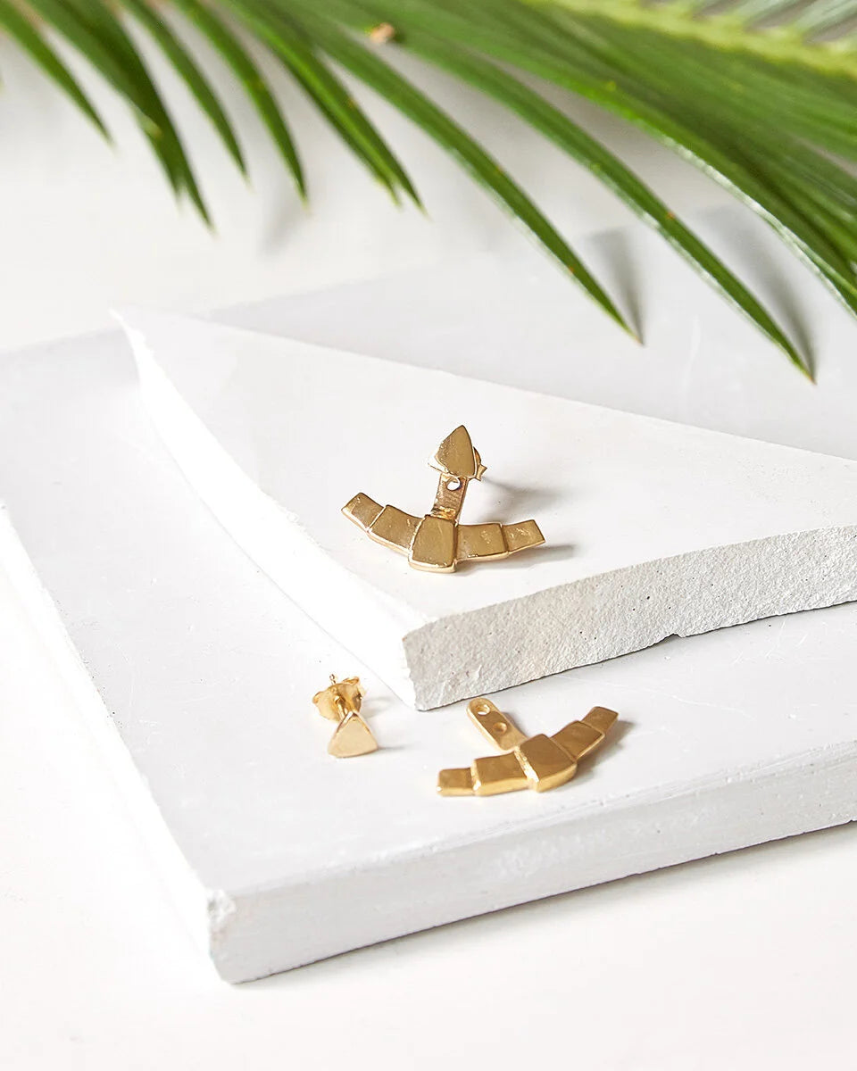 Gold-toned geometric fan ear jacket earrings displayed on a white textured block with green palm leaves in the background.