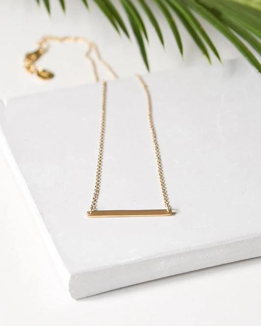 A delicate gold foundation bar necklace with a matching chain and clasp, displayed on a white textured block with green palm leaves in the background.