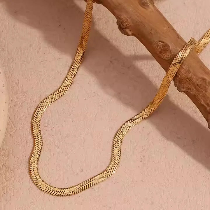 A gold wave snake chain necklace draped over a textured beige surface and a rustic wooden branch.