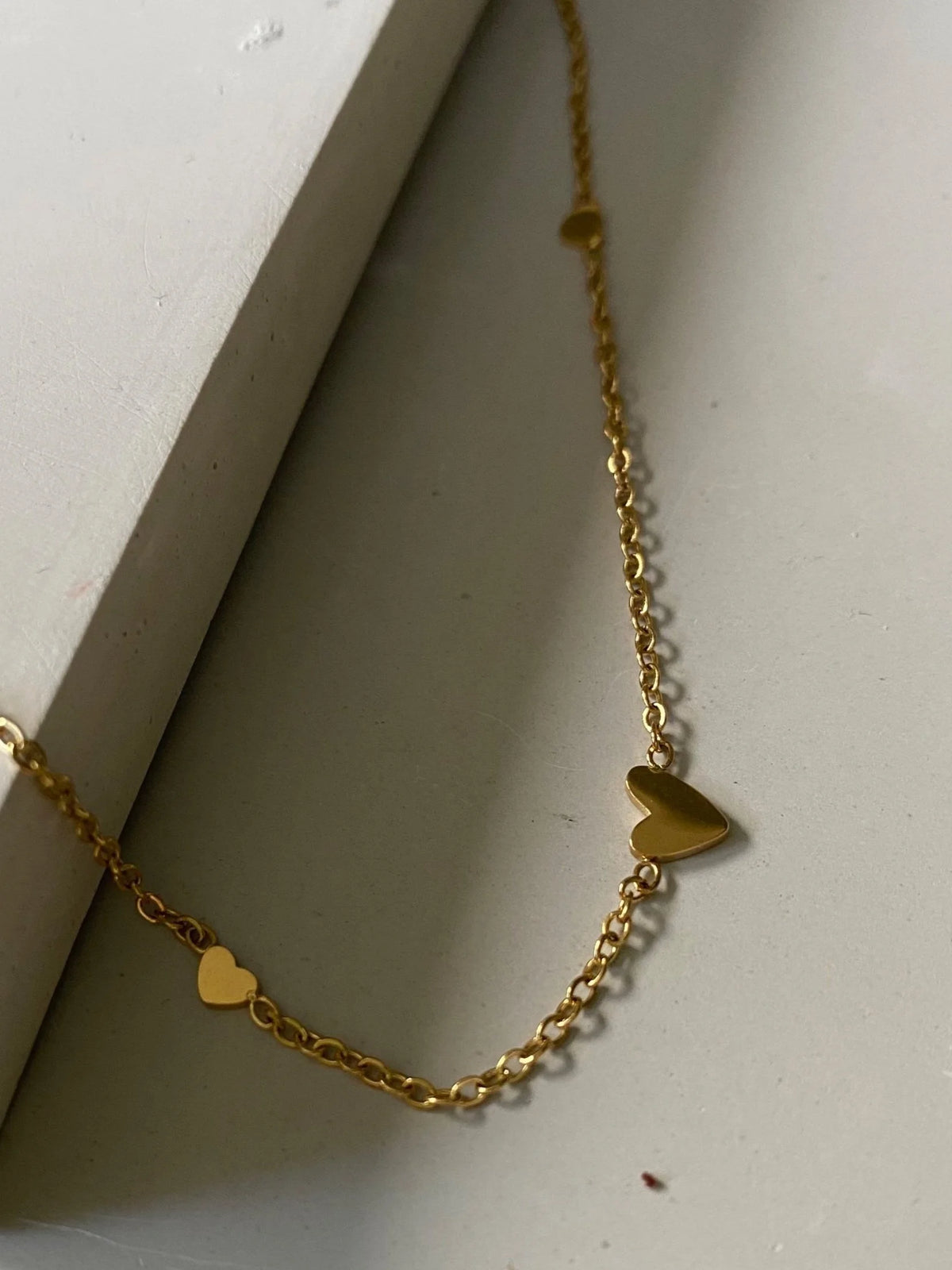 A delicate gold chain necklace with three small heart charms, displayed on a neutral light gray surface.