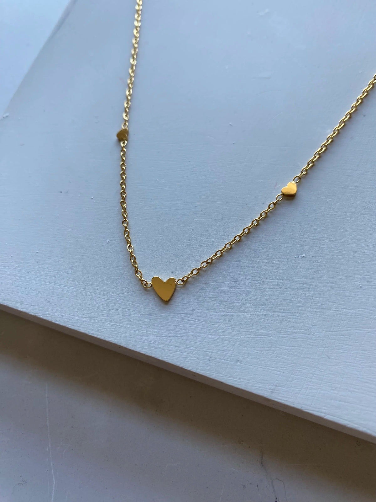 A delicate gold chain necklace featuring three small heart charms in varying sizes, displayed on a light gray textured surface.