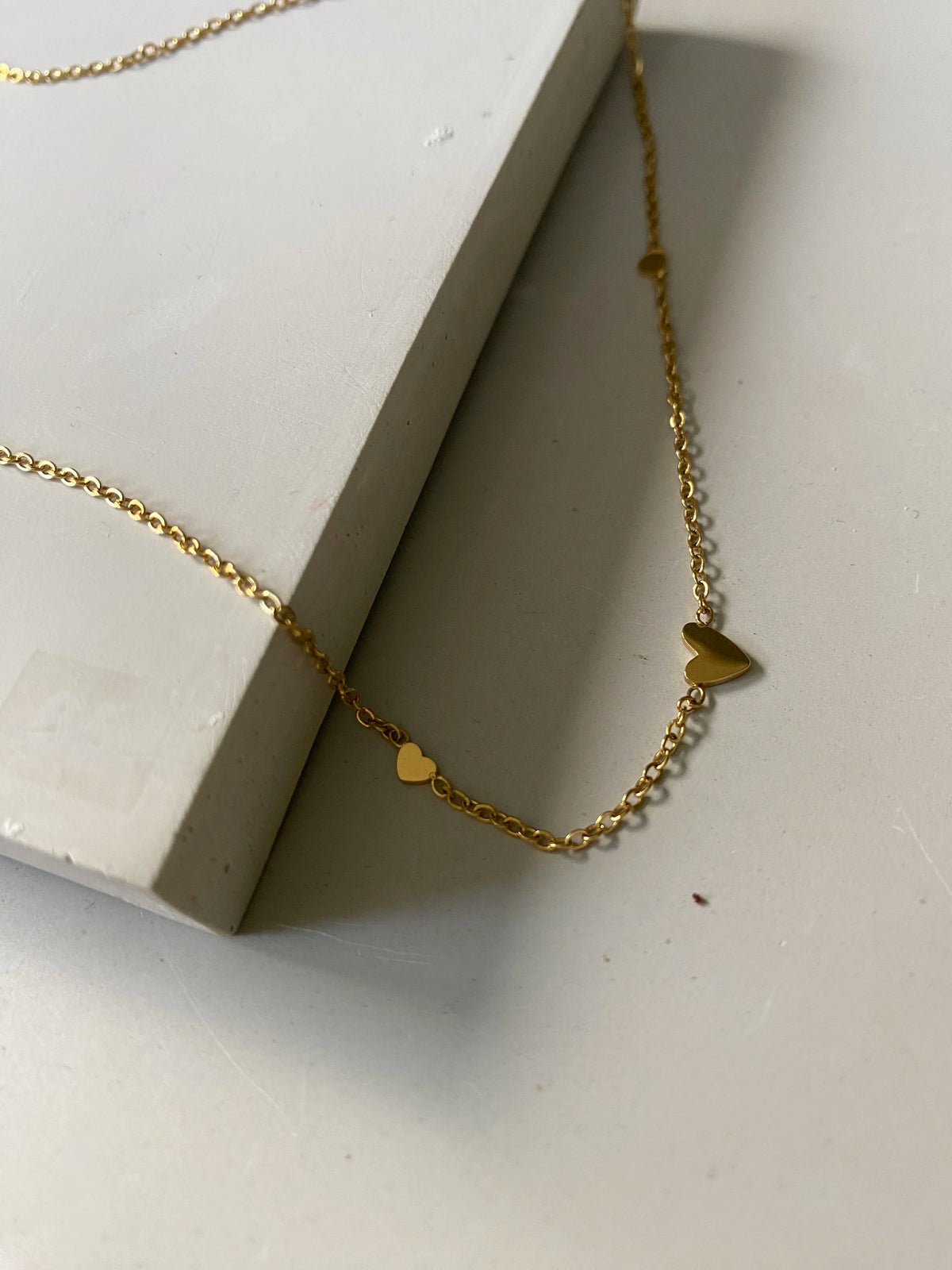 A delicate gold chain necklace with three small heart charms, draped over a white rectangular block on a light gray surface.
