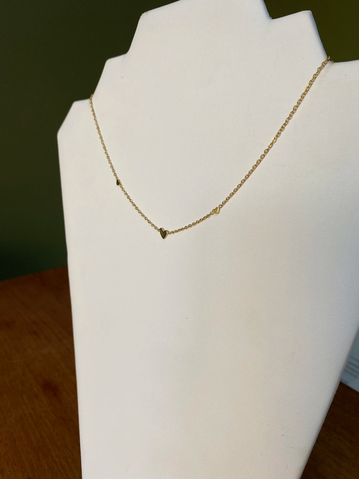 A delicate gold chain necklace with small heart charms displayed on a white jewellery bust against a dark green background.