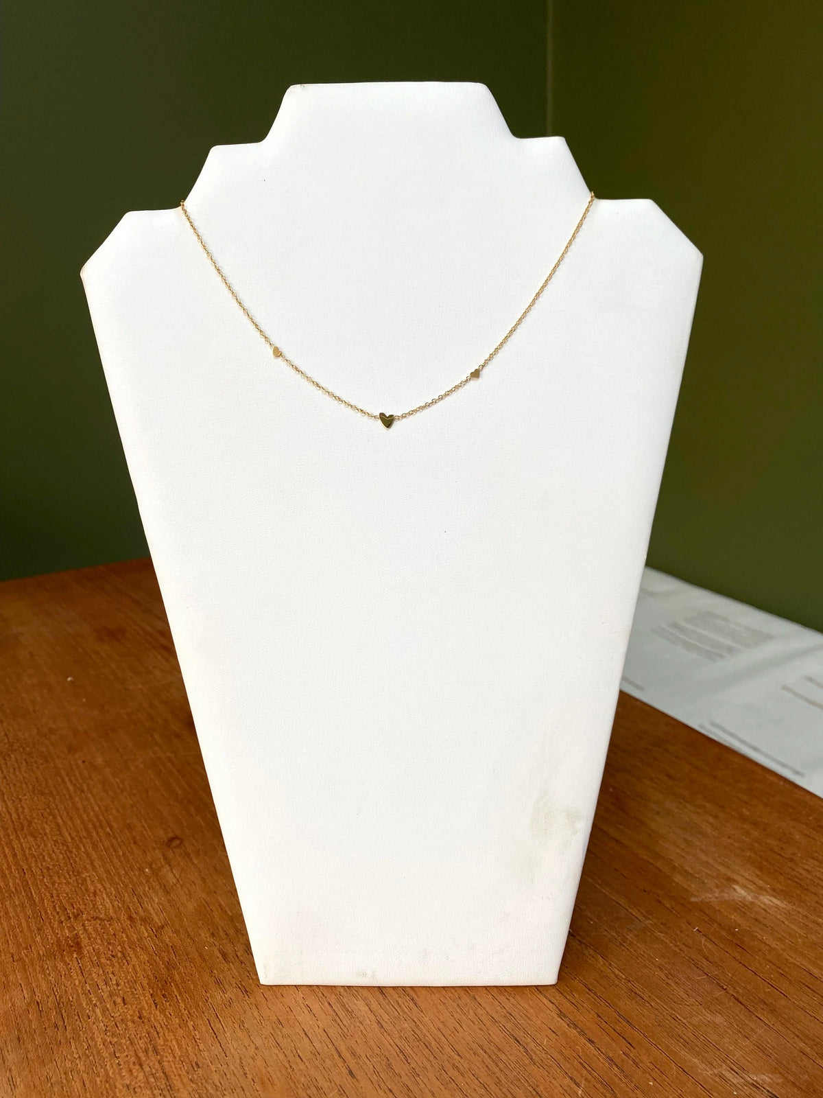 A delicate gold necklace with a small heart pendants displayed on a white jewellery bust, set on a wooden table against a dark green background.
