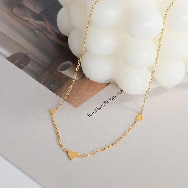 A delicate gold chain necklace with three small heart charms, styled on a white surface next to a white bubble candle.