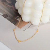 A delicate gold chain necklace with three small heart charms, styled on a white surface next to a white bubble candle.