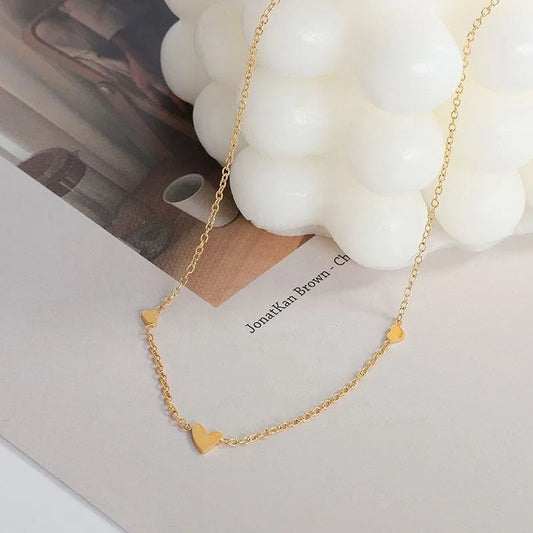 A delicate gold chain necklace with three small heart charms, styled on a white surface next to a white bubble candle.