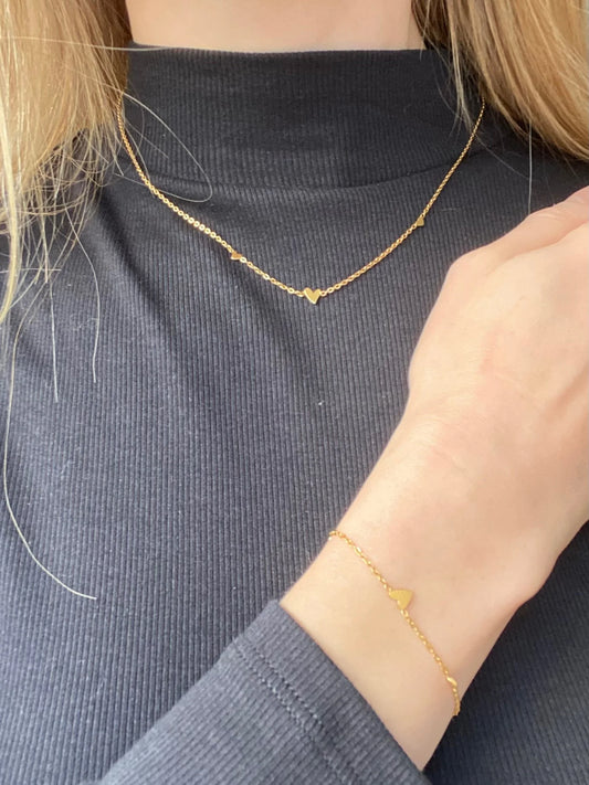 A close-up of a person wearing a delicate gold necklace and matching gold bracelet, both featuring a small heart-shaped charm, against a dark gray ribbed turtleneck sweater.