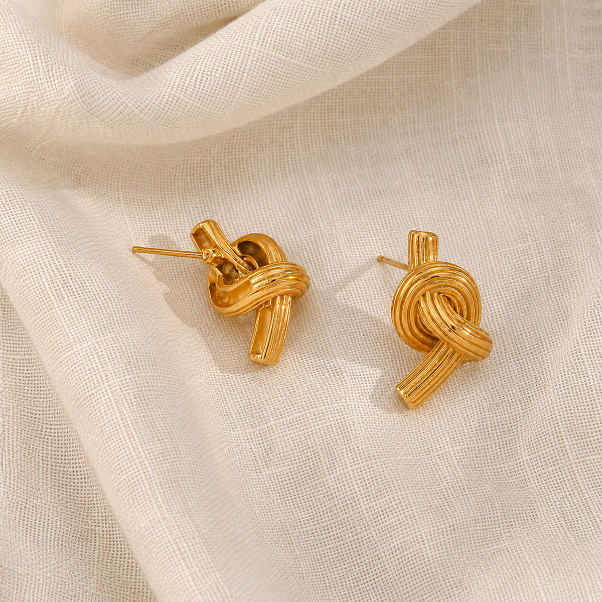 A pair of gold knot stud earrings displayed on a textured, draped cream-colored fabric background. One earring is turned over to see the back and the post 
