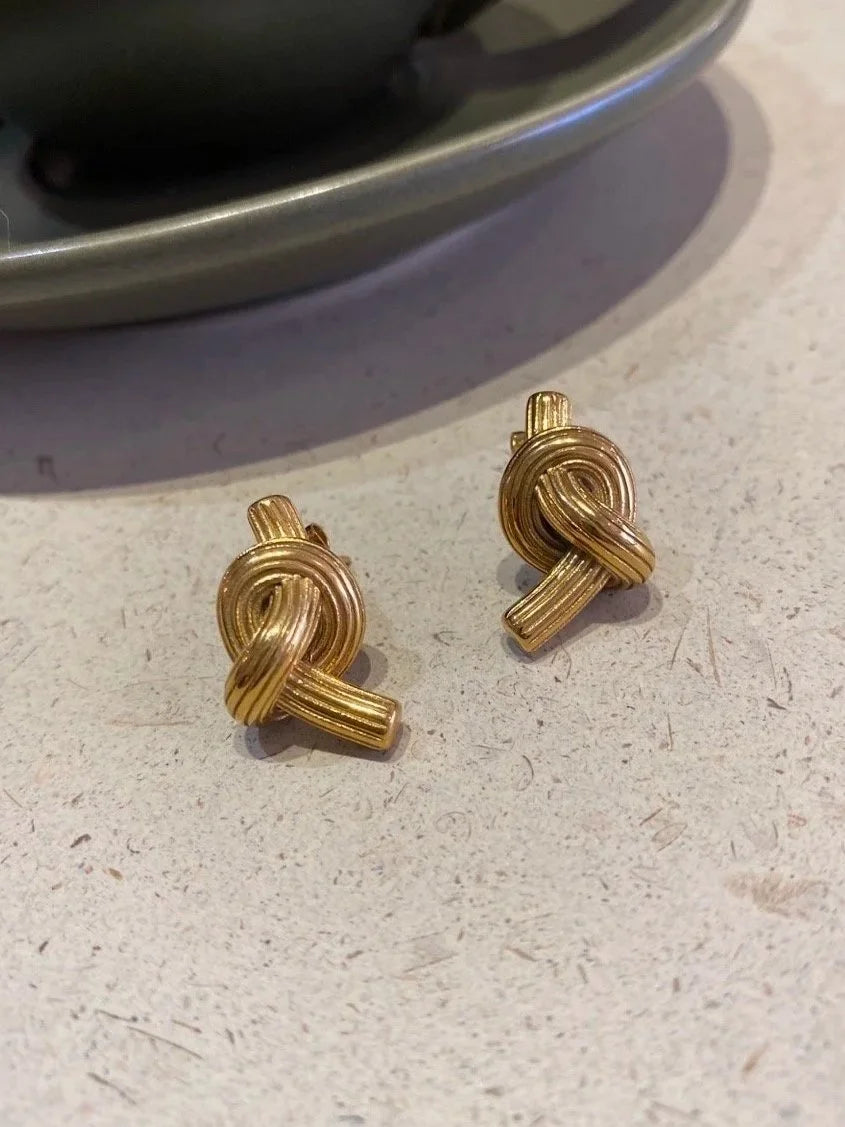 A pair of gold knot stud earrings resting on a light speckled countertop with a dark cup and saucer blurred in the background.