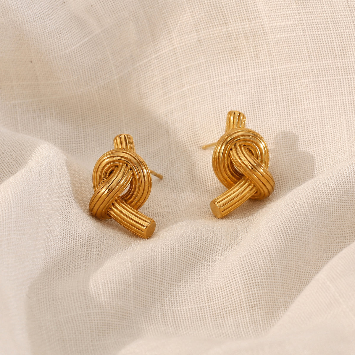 A pair of gold knot stud earrings resting on draped off-white linen fabric.