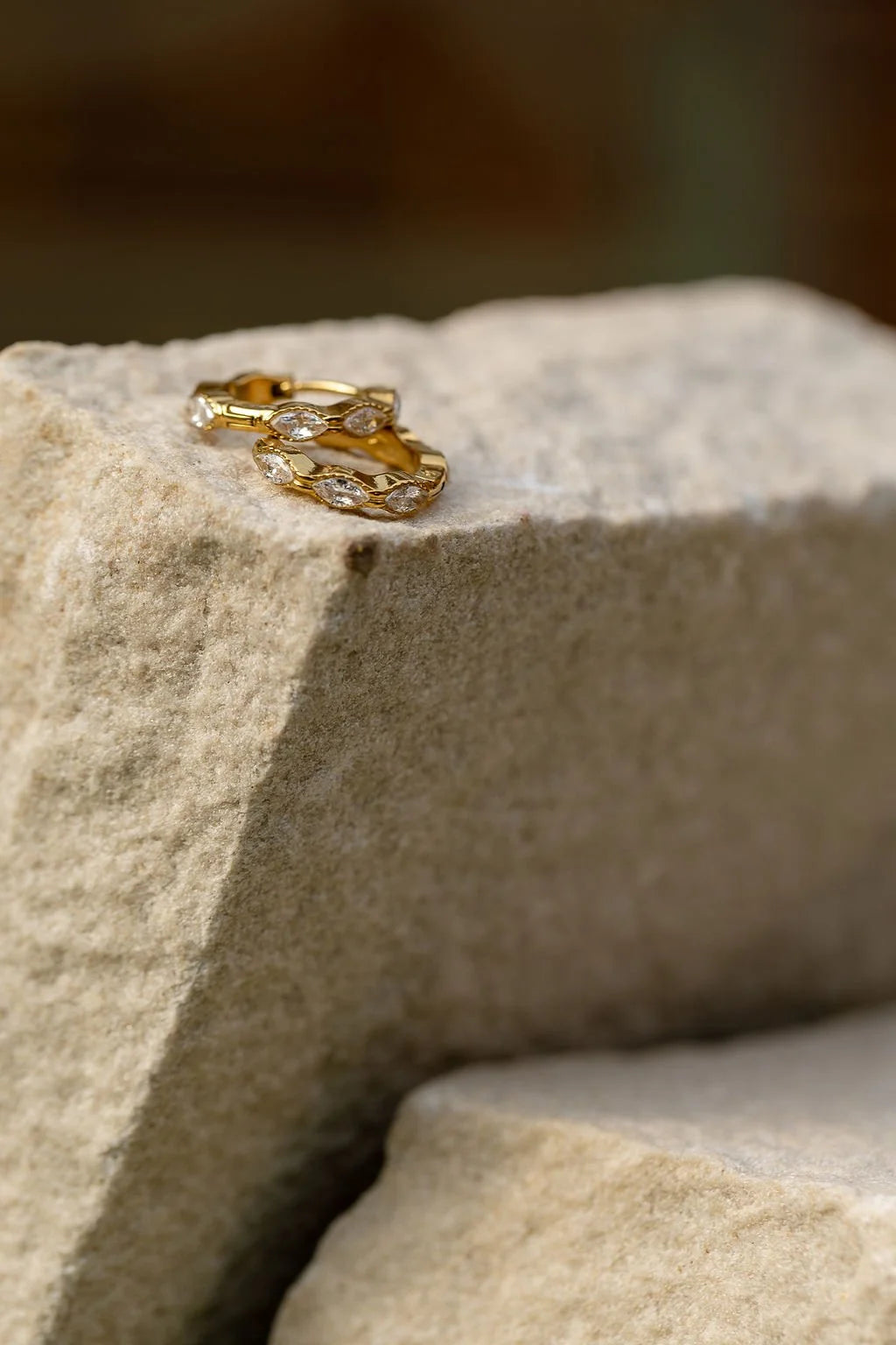 A pair of gold Luna Huggies with clear cubic zirconia crystals, resting on a textured light beige stone block.