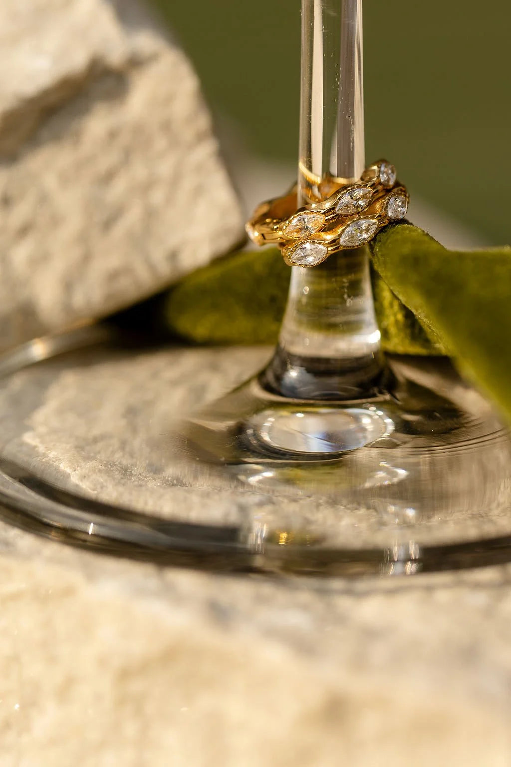 A close-up of gold Luna huggie hoops with clear cubic zirconia crystals, resting on the stem of a clear glass or wine glass, with green velvet and light stone in the background.