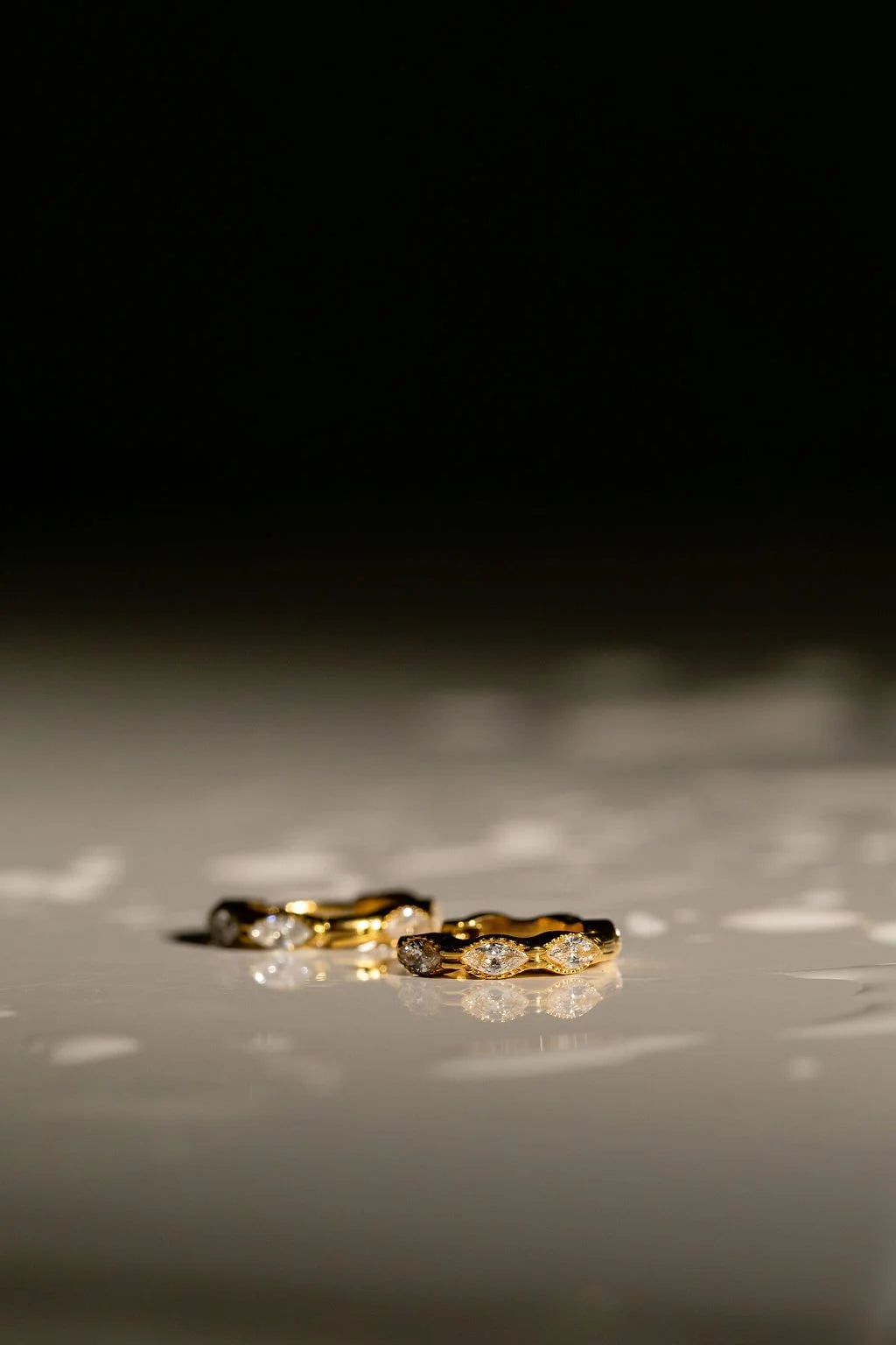 A pair of Luna Huggie hoops with small clear cubic zirconia crystals, photographed on a glossy light surface against a dark background with water droplets around.