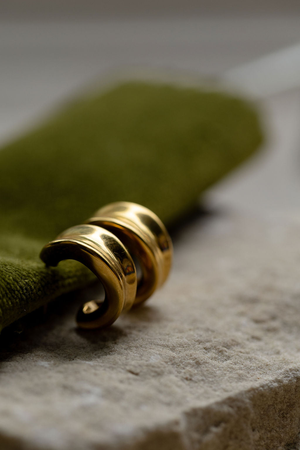 A pair of polished gold Sculpted hoop earrings resting on a textured stone surface next to a folded olive green fabric.