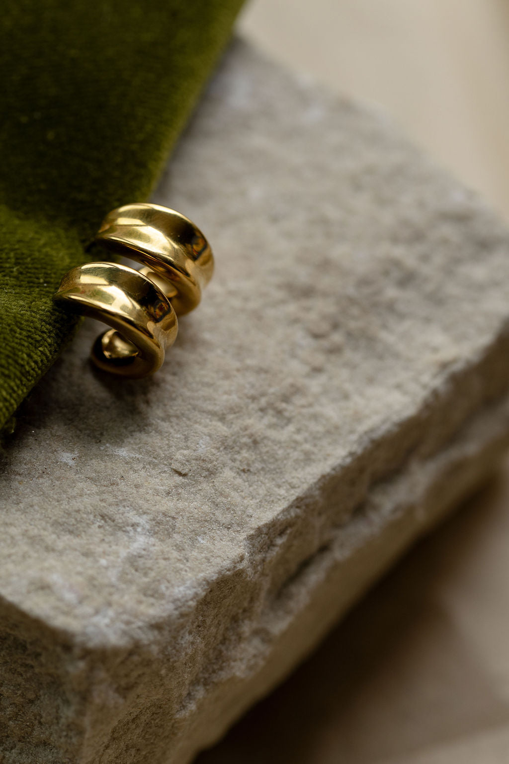 A pair of polished gold chunky hoop earrings resting on a textured light grey stone slab attached to an olive green fabric accent.