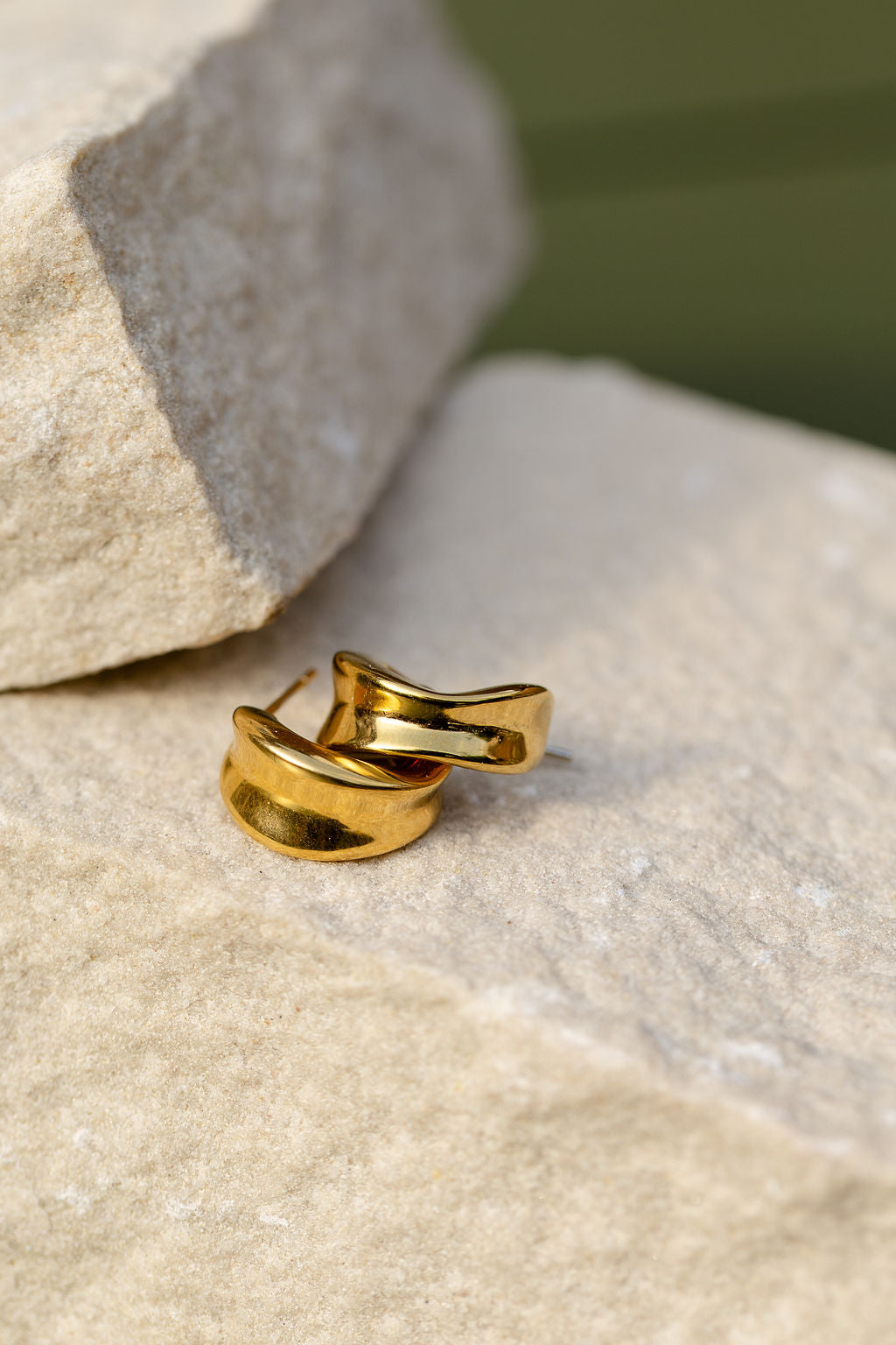 A pair of polished gold sculpted hoop earrings resting on a textured beige stone surface with a blurred green background.