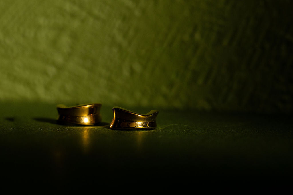 Two gold tactile sculpted hoops resting on a dark surface against a textured olive-green background.