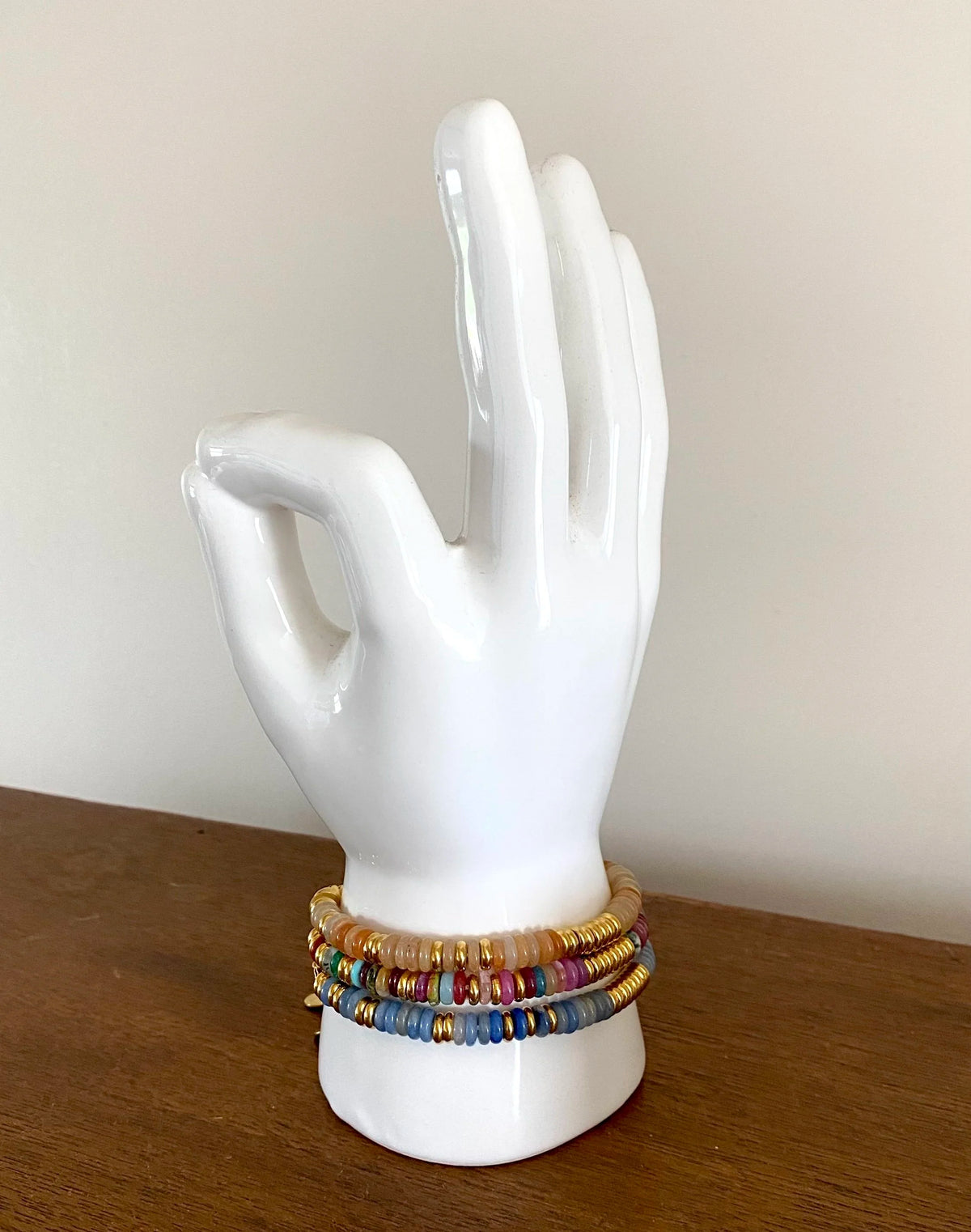 A set of three colourful beaded bracelets displayed on a white ceramic hand-shaped jewellery stand, resting on a wooden surface against a plain beige wall.