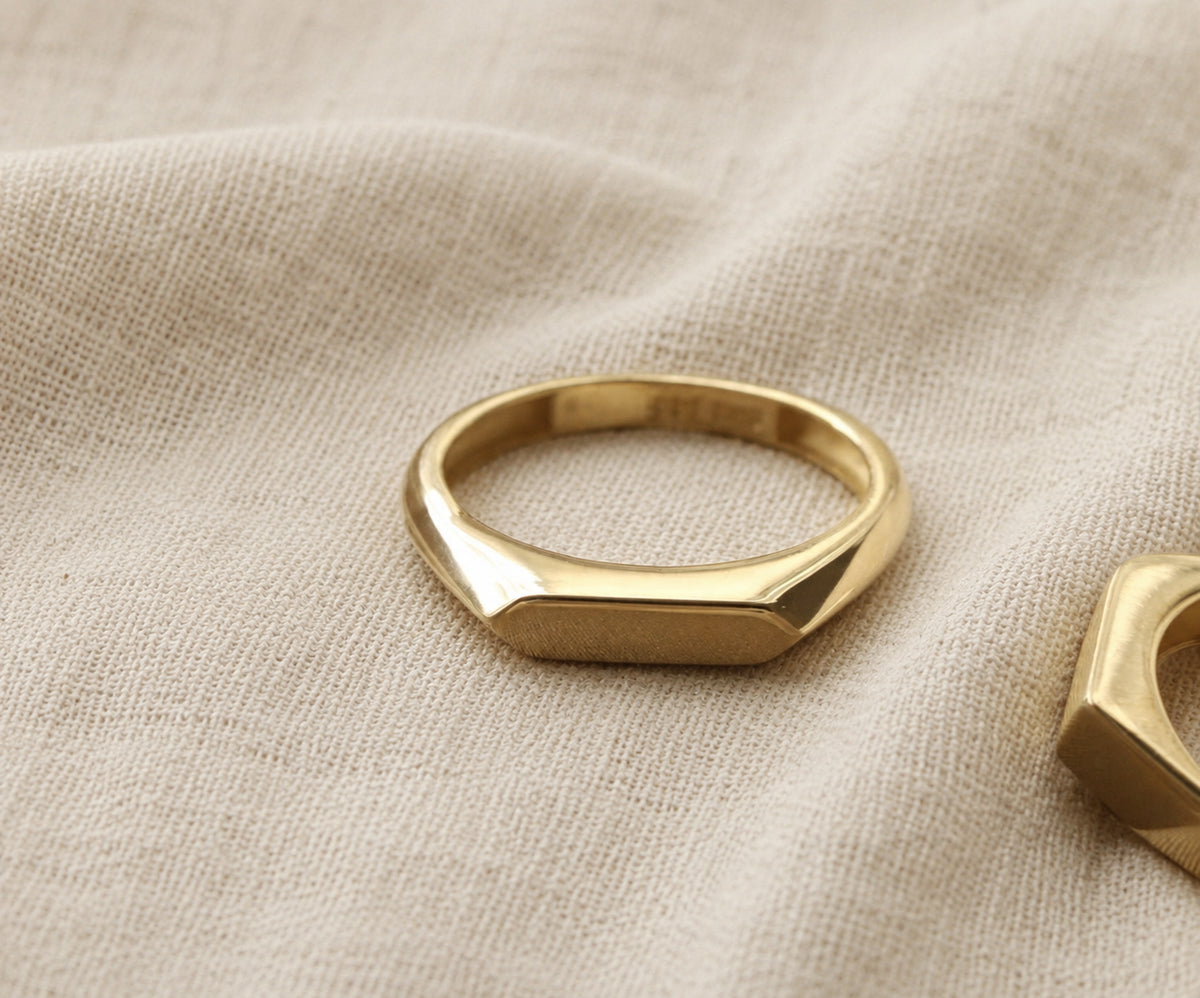 A gold-toned slim signet ring with a flat, faceted rectangular face, resting on a textured beige linen fabric.