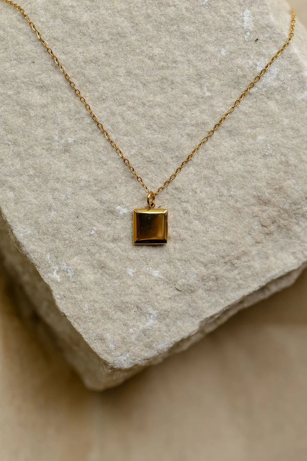 A delicate gold chain necklace with a small square pendant, displayed on a textured beige stone block.