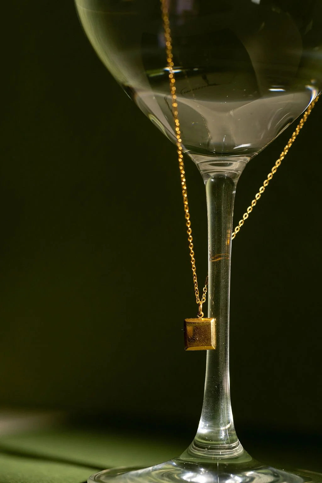 A gold necklace with a small square pendant hangs around the stem of a clear wine glass against a dark olive green background.