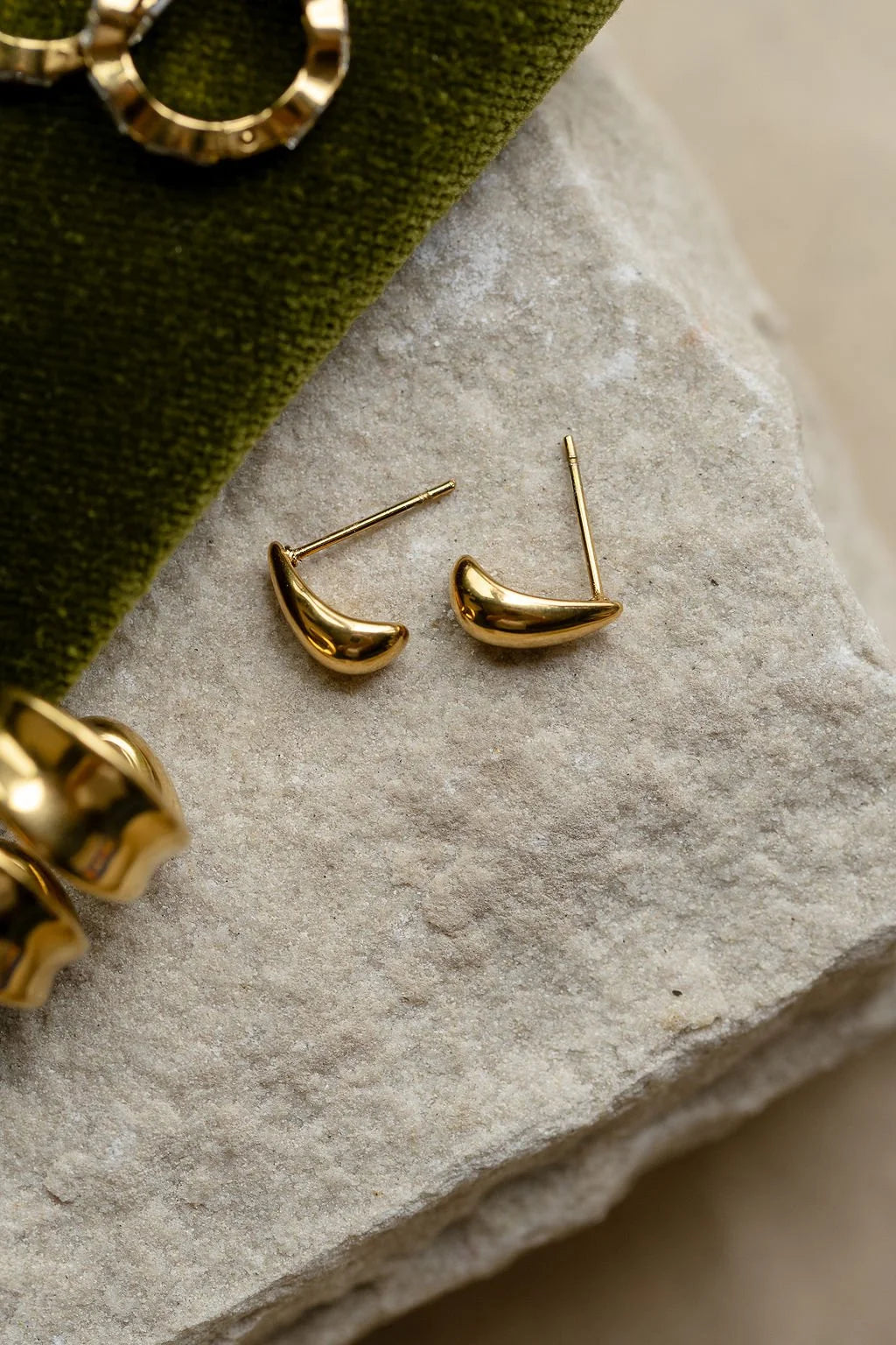 A pair of small gold crescent-shaped stud earrings resting on a textured light gray stone surface, with a dark green velvet fabric and partial gold jewelry visible around them.