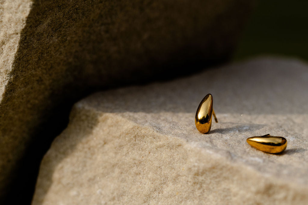 Two small gold teardrop-shaped stud earrings resting on a textured light stone surface, with a darker stone block in the background.