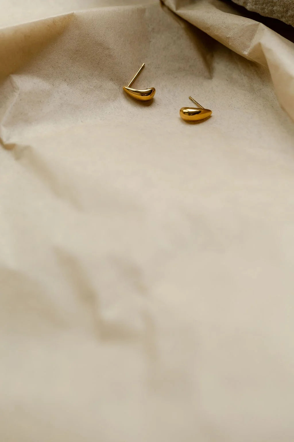 A pair of small gold teardrop shaped stud earrings resting on crumpled beige paper.