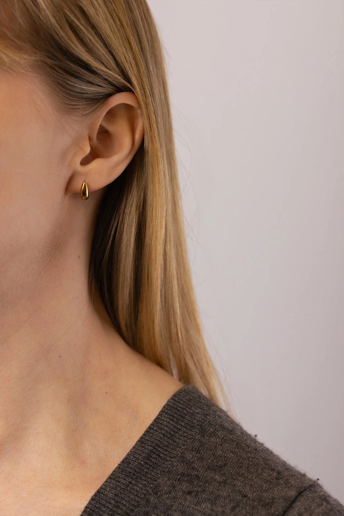 A close-up of a model's ear wearing a small gold teardrop stud earring, with her blonde hair and shoulder visible against a plain light background.