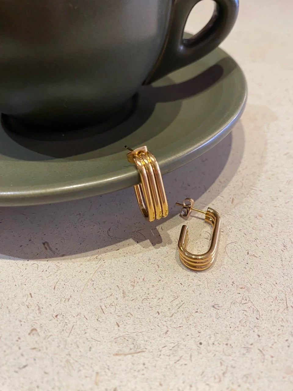 A pair of gold-toned geometric Trio hoop earrings resting on a light speckled countertop next to a dark green ceramic cup and saucer.