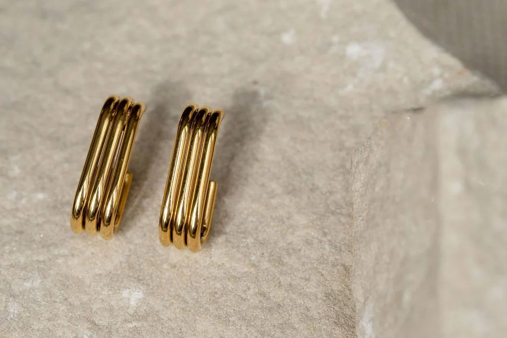 A pair of gold-plated Trio hoop earrings resting on a textured light beige stone surface.