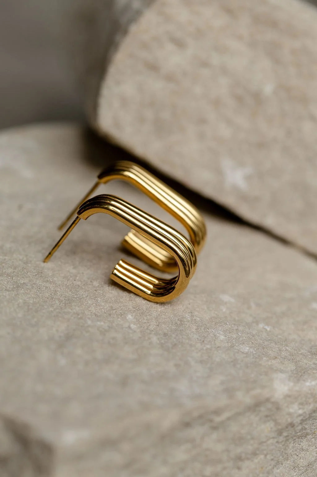 A gold-toned geometric earring with a three stands formed into a rectangular hoops, resting on a textured stone surface.