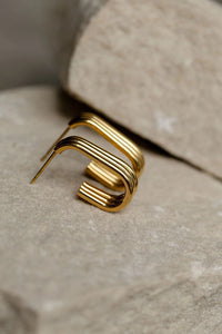 A gold-toned geometric earring with a three stands formed into a rectangular hoops, resting on a textured stone surface.