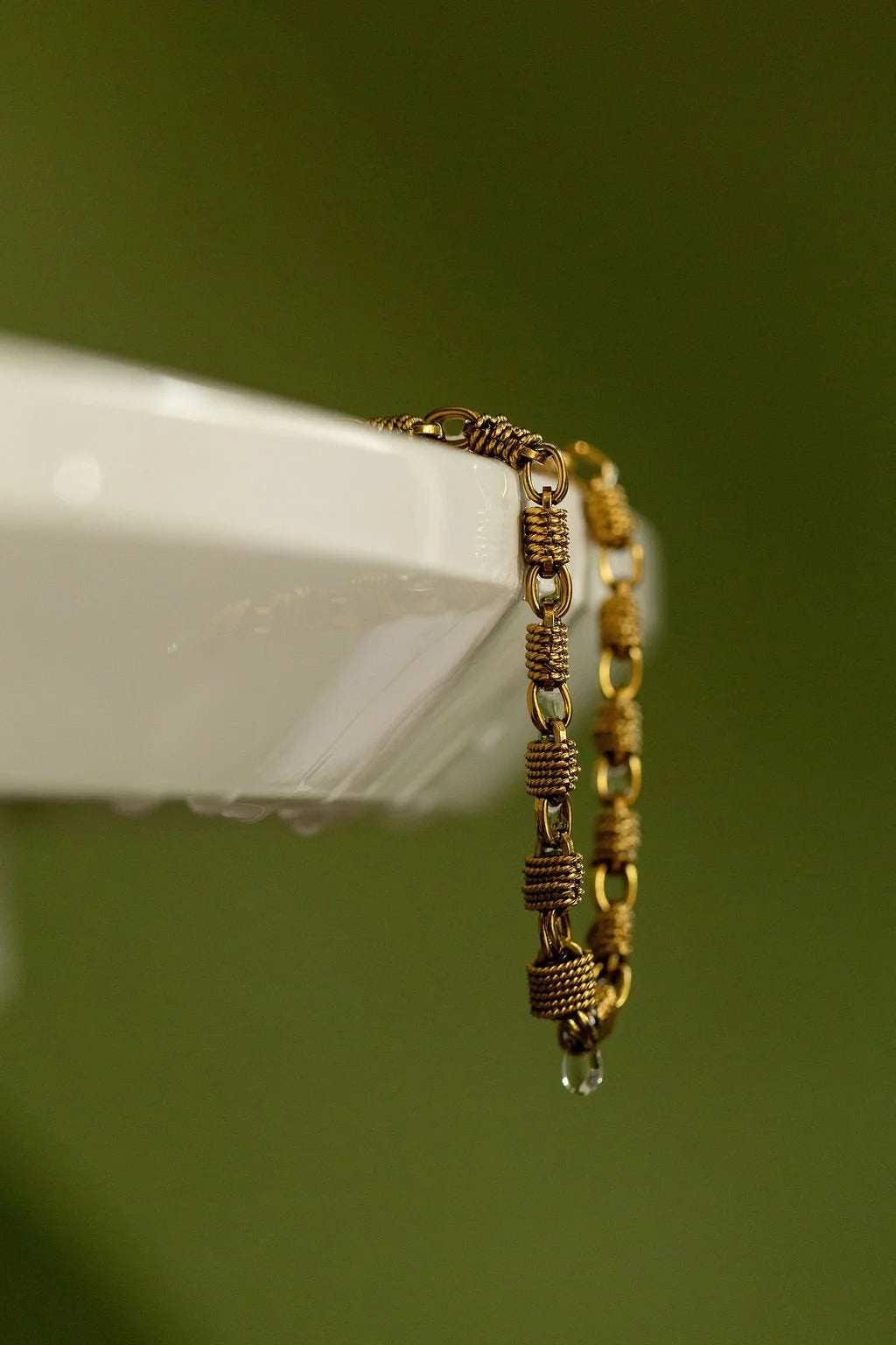 A close-up of a gold-toned Woven Chunky chain necklace draped over the edge of a white surface, with a single clear water droplet hanging from the lowest link, against a soft olive green background.
