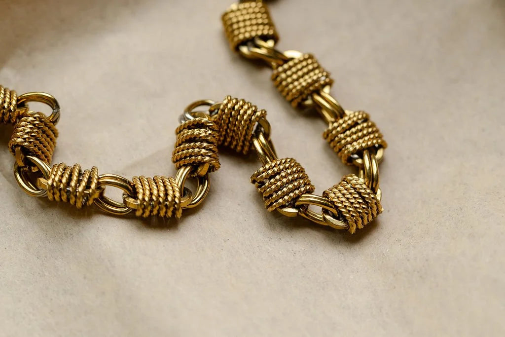 A close-up of a gold-toned Woven Chunky chain necklace with textured, woven cylindrical links resting on a neutral beige surface.