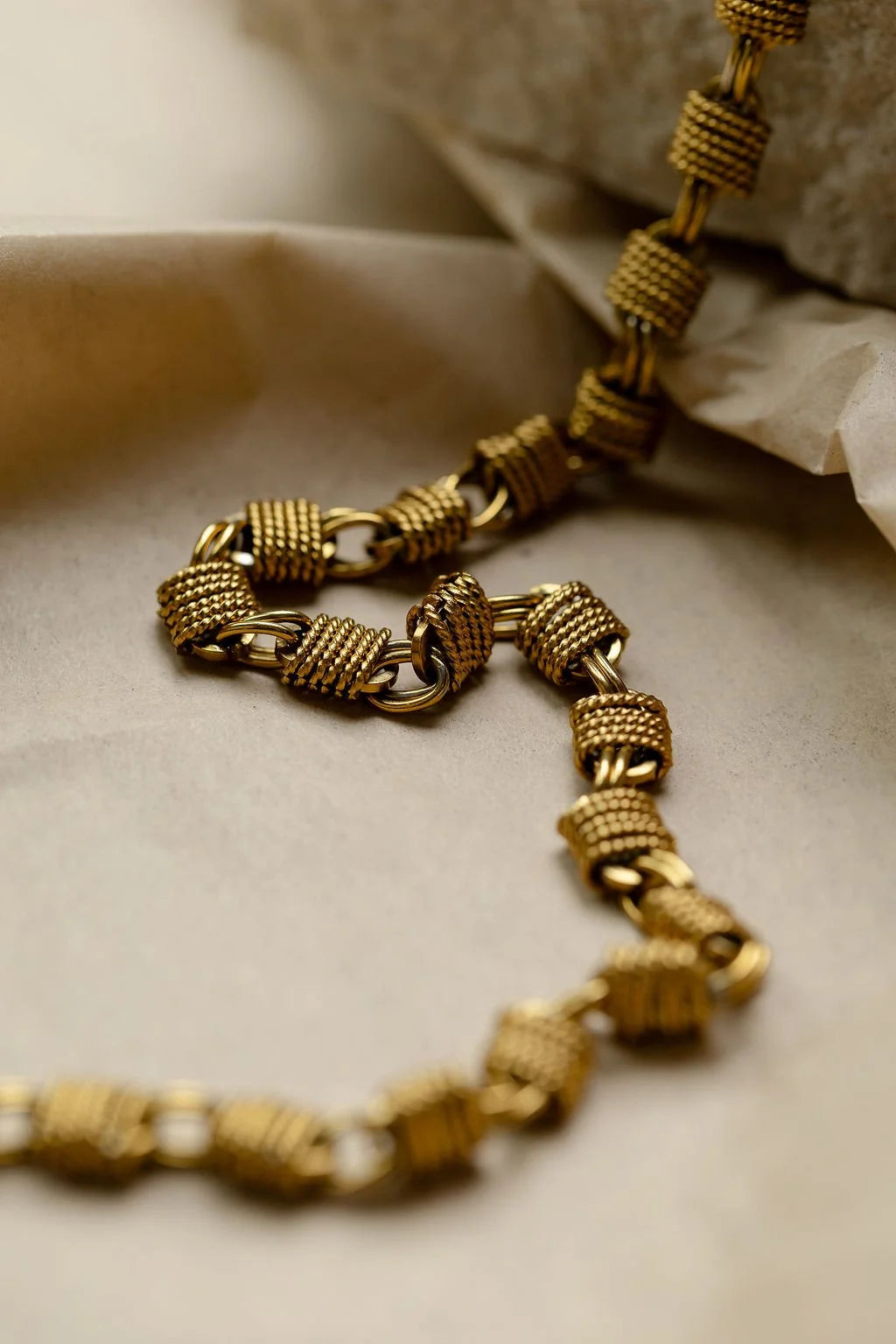 A close-up of a gold-toned Woven Chunky chain necklace with textured, woven-link segments, draped over a neutral beige surface with a soft-focus stone in the background.