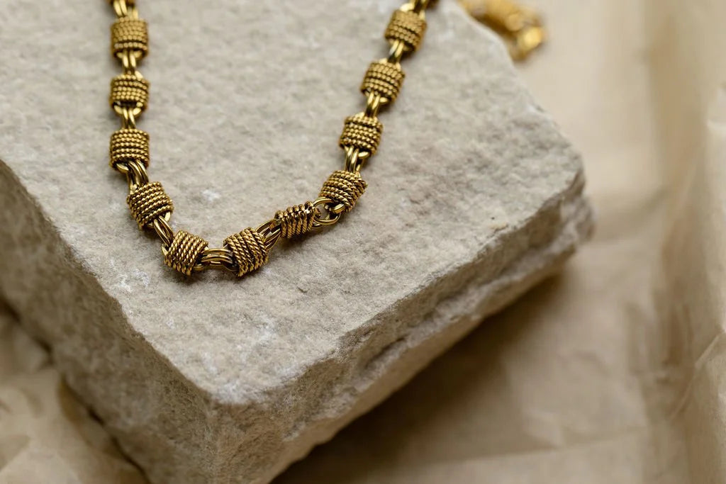 A close-up of a gold-toned chain necklace with textured, knotted cylindrical links, resting on a light gray stone block against a soft beige fabric background.